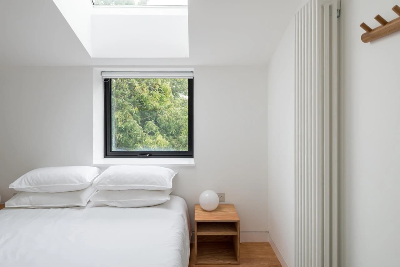 Minimalist bedroom with clean white linens and wooden nightstand positioned beneath a black-framed window overlooking lush green foliage, bathed in natural light from a skylight above.