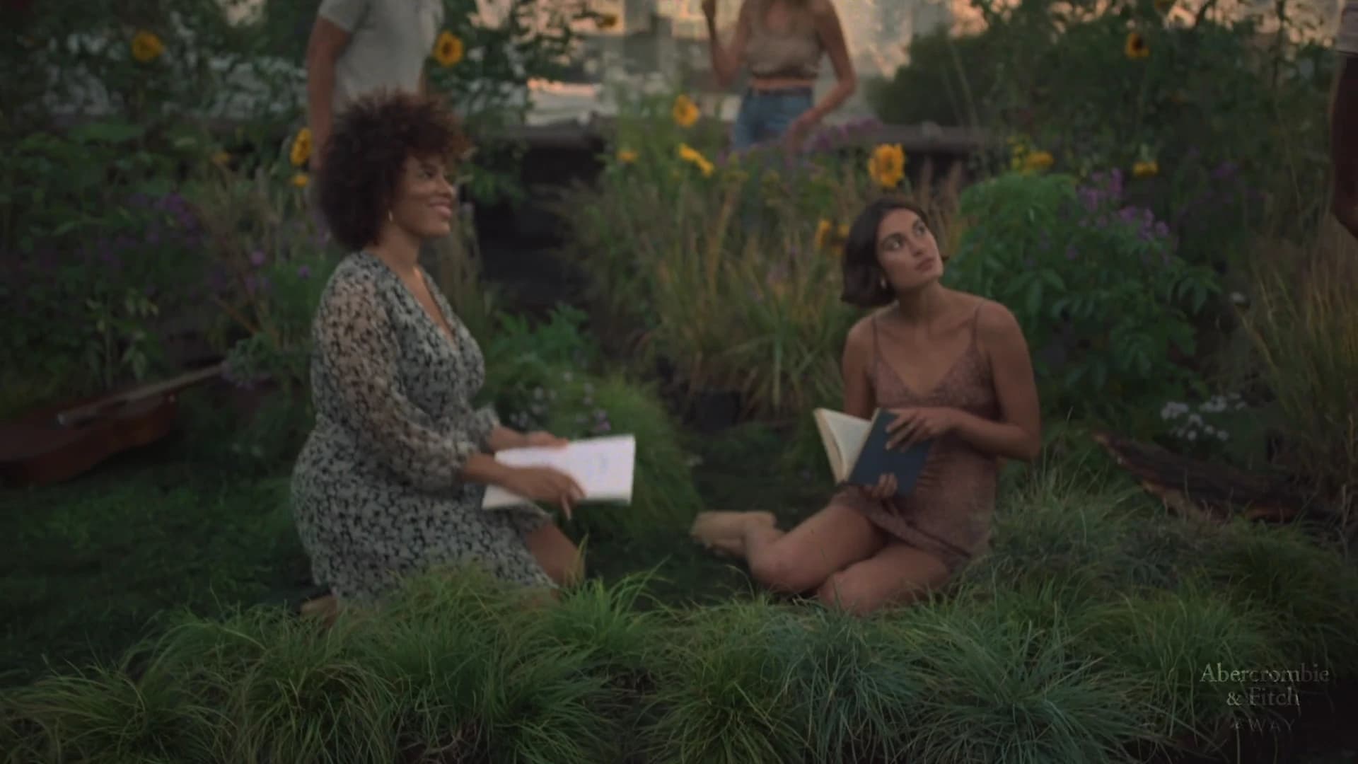 Two women engage in conversation while seated in a lush garden surrounded by ornamental grasses, sunflowers, and purple flowering plants, with softly blurred figures visible in the background among the verdant landscape.