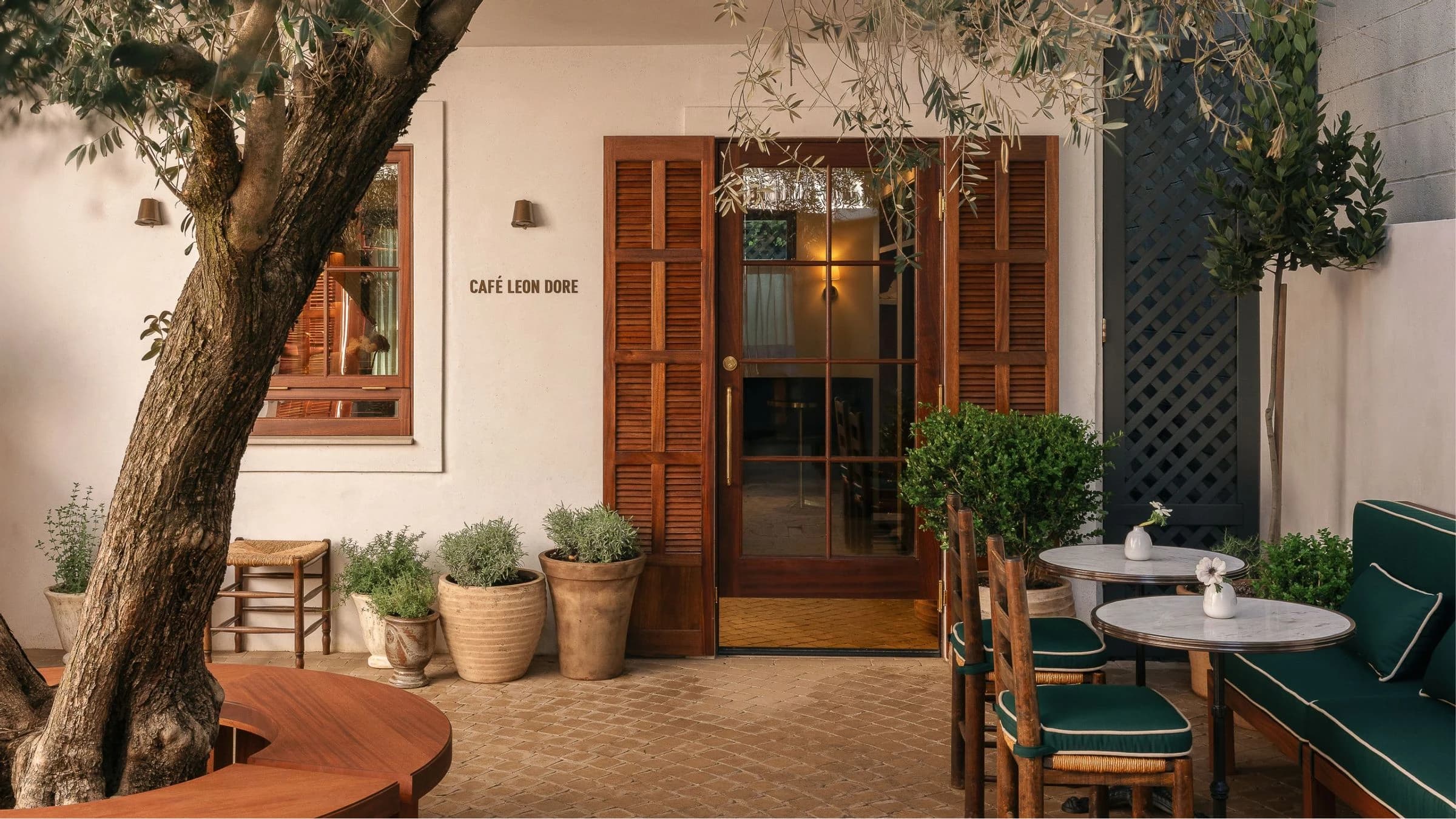 A serene Mediterranean-inspired courtyard features a centuries-old olive tree casting dappled shade over a terracotta-tiled patio, with warm wooden doors, potted herbs and plants, and intimate seating areas appointed with green cushioned chairs and marble-topped tables.